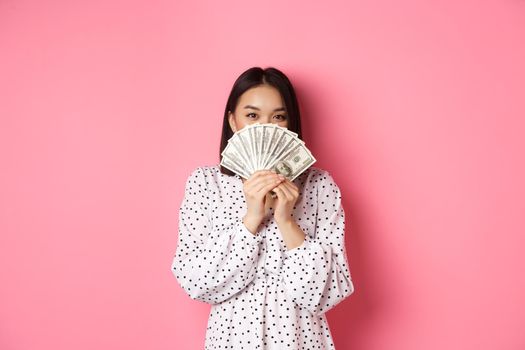 Shopping concept. Cute asian woman hiding face behind money dollars, peeking at camera, standing over pink background.