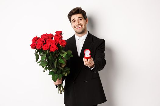 Handsome smiling man in black suit, holding roses and engagement ring, making a proposal to marry him, standing against white background.