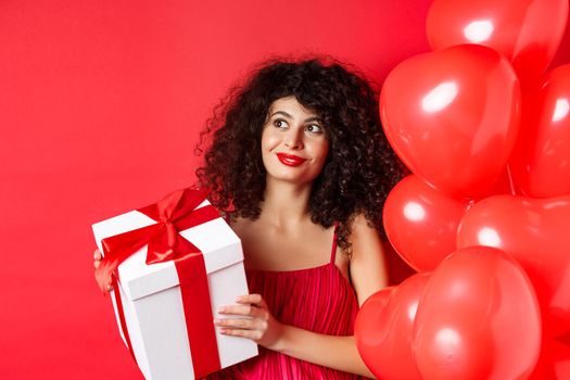Gorgeous woman with makeup and evening dress, receive surprise gift and smiling, standing on red background near Valentines day romantic balloons.