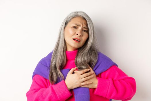 Worried asian mother holding hands on heart and grimacing, looking anxious and having pain in chest, feeling hurt, standing over white background.