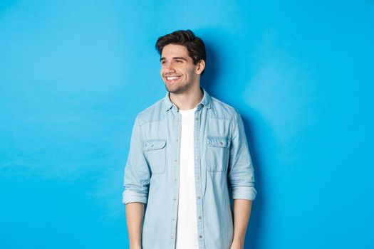 Handsome smiling adult man in casual outfit, smiling and looking left at promo offer, standing against blue background.