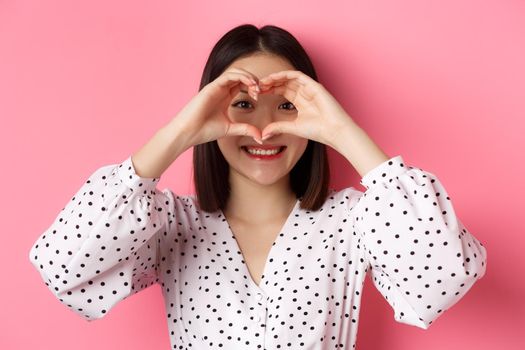 Beauty and lifestyle concept. Close-up of lovely asian woman showing heart sign, smiling and feeling romantic on valentines day, standing over pink background.