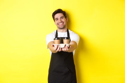 Friendly barista in black apron giving takeaway order, holding two cups of coffee and smiling, standing over yellow background.