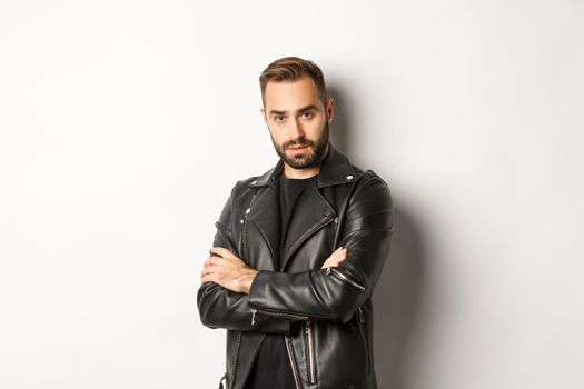 Attractive macho man with beard, wearing leather jacket smiling, looking confident with hands crossed on chest, white background.