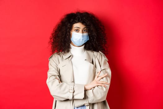 Covid-19, social distancing and quarantine concept. Portrait of skeptical girl in face mask and trench coat, cross arms on chest and staring unamused at camera, red background.