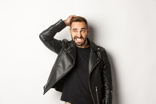 Confused macho guy in black leather jacket looking awkward and unsure, scratching head puzzled and stare left, white background.