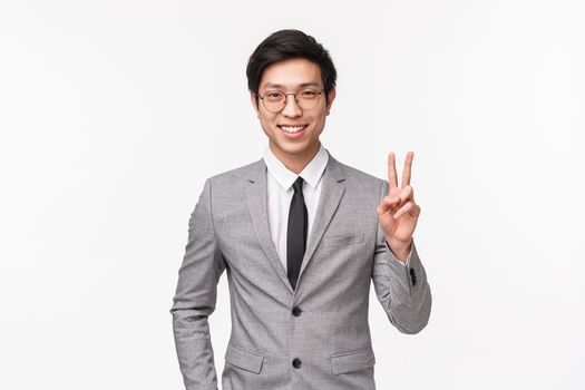 Portrait of successful determined and confident young asian man feeling he can get the deal for enterprise, standing in gray suit, showing peace sign and smiling, standing white background.