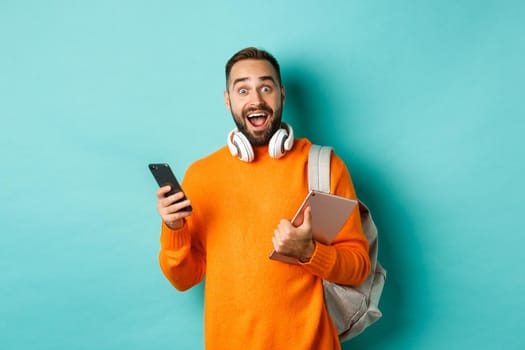 Handsome man student with headphones and backpack, holding digital tablet and smartphone, looking amazed at camera, standing against turquoise background.