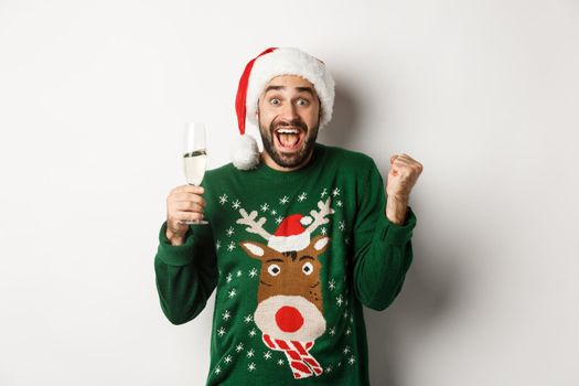 Christmas party and holidays concept. Excited man in Santa hat celebrating New Year, drinking champagne and rejoicing, standing over white background.