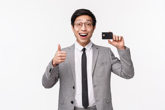 Waist-up portrait of excited, happy smiling asian male office worker, employee in grey suit and glasses, showing credit card and make thumbs-up sign, recommend bank, white background.