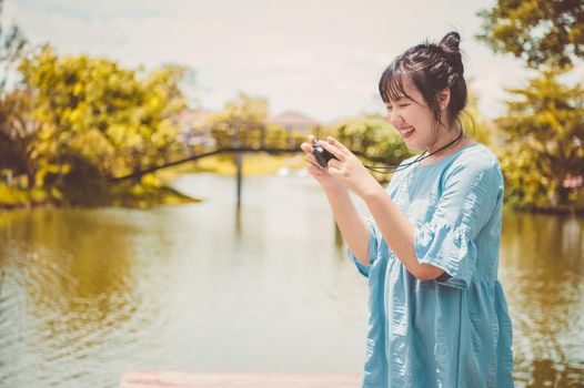 Asian woman in blue dress in public park carrying digital mirrorless camera and taking photo without facial mask in happy mood. People lifestyle and leisure concept. Outdoor travel and Nature theme.