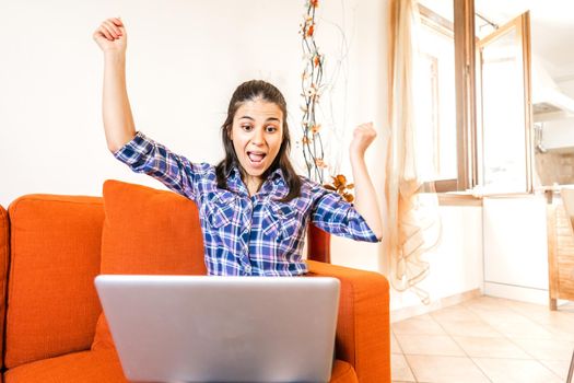 Brunette smart gen z girl rejoices in front of with open raised arms sitting at home sofa. Shocked young woman looking at computer screen in living room. Happy student looks results of a school exam