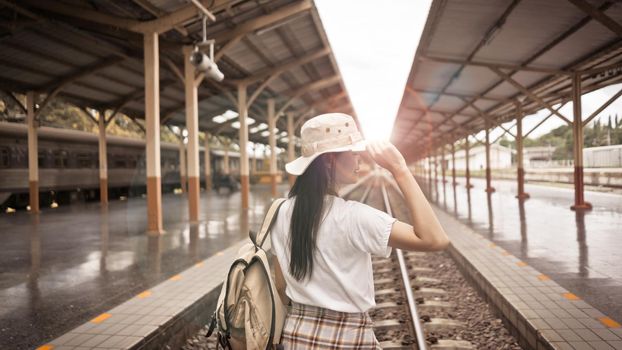 Young female tourist good feeling with a backpack and holding her hat on the railroad tracks at the train station in the sunset. Happy Holiday and Freedom active lifestyle concept.