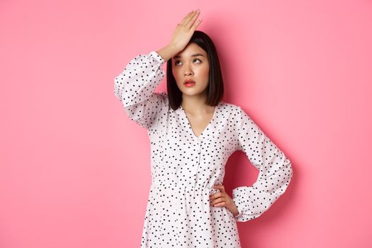 Annoyed and tired asian woman making facepalm, slap forehad and roll eyes disappointed, standing in white dress against pink background.