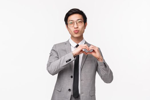 Waist-up portrait of handsome romantic male office worker in grey suit, showing heart sign and fold lips in kiss, looking camera, express like or sympathy, standing white background.