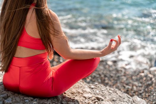 A young woman in red leggings and a red top with long loose hair practices yoga outdoors by the sea on a sunny day. Women's yoga, fitness, Pilates. The concept of a healthy lifestyle, harmony