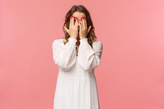 Portrait of silly cute girl with blond short hair, wear white dress, hiding face behind hands, promise to wait for signal but peeking through fingers, cant resist temptation, stand pink background.