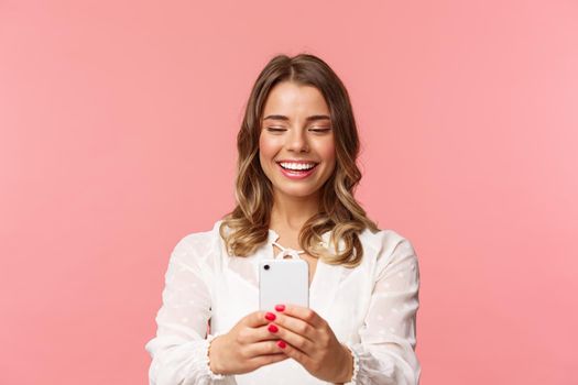 Close-up portrait of happy charming caucasian woman with short fair hair, white dress, record video of concert on mobile phone, taking photo using smartphone, standing pink background.