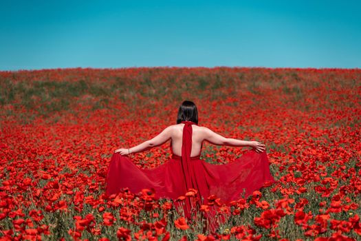 Young woman stands with her back in a long red dress and hat, posing on a large field of red poppies.