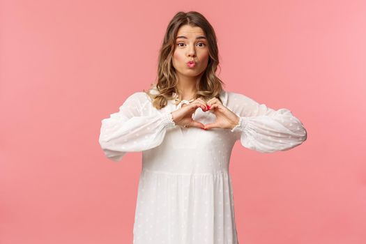 Beauty, fashion and women concept. Portrait of tender lovely young blond girl in beautiful white dress, fold lips in kiss and make heart sign near chest, standing pink background, show sympathy.