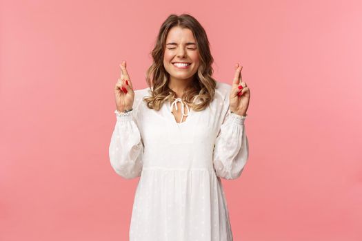 Portrait of hopeful lucky cute blond girl in white dress praying, anticiapte miracle, making wish to win, cross fingers good luck, smiling and close eyes and pleading god, pink background.