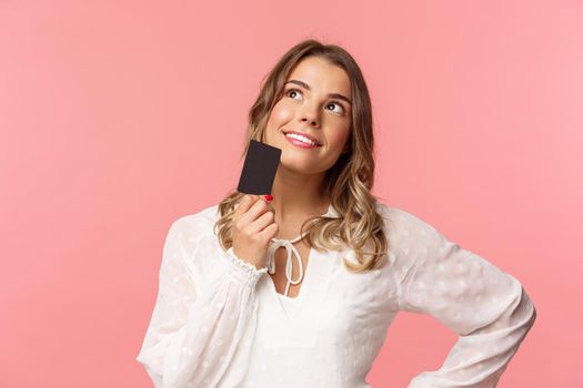 Close-up portrait of thoughtful and creative blond woman in white dress, touching chin with credit card and smiling dreamy as looking up and thinking, picturing what buy as present, pink background.