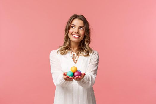 Holidays, spring and party concept. Portrait of dreamy good-looking blond girl in white dress, holding painted eggs, celebrating Easter, orthodox holy day, smiling and looking away.