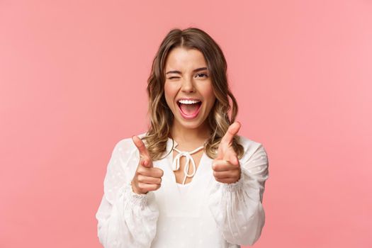 Close-up portrait of sassy and carefree good-looking emotive blond girl, pointing finger pistols at camera as asking join her team, become member, inviting try product yourself, smiling cheeky.