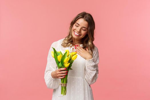 Holidays, beauty and spring concept. Portrait of pleased, cute romantic blond girl receive beautiful bouquet of flowers, looking at yellow tulips and touch heart grateful, smiling.