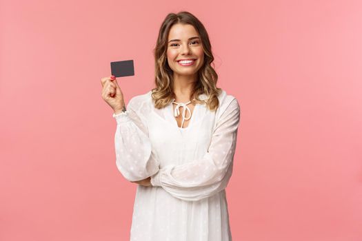 Portrait of pleased good-looking blond european female in white dress, show credit card with satisfied expression, smiling camera, recommend bank services, use payment online, pink background.