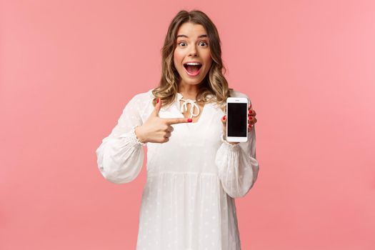 Portrait of impressed, excited young blond woman showing something awesome on display, pointing mobile phone screen and smiling astonished, brag with her recent match on dating app.