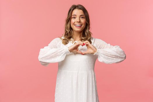 Beauty, fashion and women concept. Romantic passionate blond european female in white cute dress, make heart sign near chest and smiling at camera excited, show sympathy.