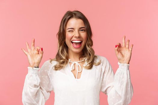 Close-up portrait of beautiful blond girl in white dress guarantee you will enjoy this event, show okay sign, confirm everything excellent, wink and smiling carefree, standing pink background.