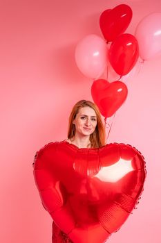 Beautiful redhead girl with red heart baloon posing. Happy Valentine's Day concept. Studio photo of beautiful ginger girl dancing on pink background.