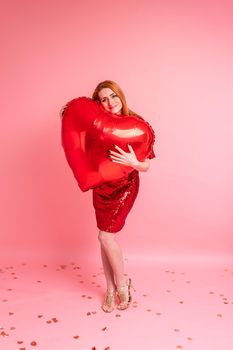 Beautiful redhead girl with red heart baloon posing. Happy Valentine's Day concept. Studio photo of beautiful ginger girl dancing on pink background.