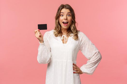 Portrait of happy excited good-looking blond girl in white dress, being impressed and pleased with cool banking service, showing credit card, smiling camera, standing pink background.