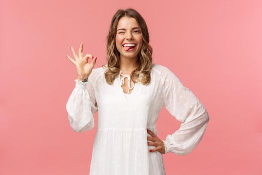 Portrait of happy smiling blond woman with short curly haircut, show tongue and okay sign, say alright, agree or approve, rate great product, guarantee quality, stand pink background.