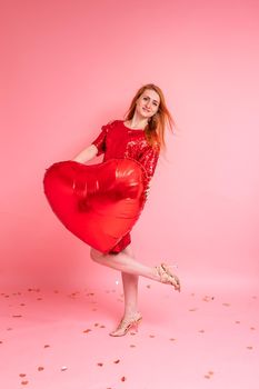 Beautiful redhead girl with red heart baloon posing. Happy Valentine's Day concept. Studio photo of beautiful ginger girl dancing on pink background.