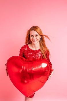 Beautiful redhead girl with red heart baloon posing. Happy Valentine's Day concept. Studio photo of beautiful ginger girl dancing on pink background.