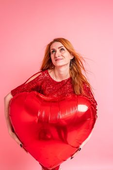 Beautiful redhead girl with red heart baloon posing. Happy Valentine's Day concept. Studio photo of beautiful ginger girl dancing on pink background.