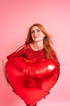 Beautiful redhead girl with red heart baloon posing. Happy Valentine's Day concept. Studio photo of beautiful ginger girl dancing on pink background.