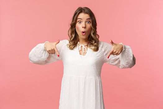 Waist-up portrait of interested, excited attractive blond 20s woman showing person something really cool, say wow pop eyes camera excited, pointing fingers down at great offer, pink background.