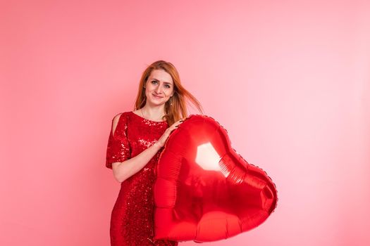 Beautiful redhead girl with red heart baloon posing. Happy Valentine's Day concept. Studio photo of beautiful ginger girl dancing on pink background.