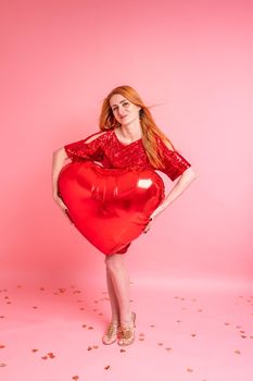 Beautiful redhead girl with red heart baloon posing. Happy Valentine's Day concept. Studio photo of beautiful ginger girl dancing on pink background.