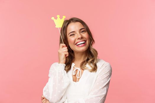 Spring, happiness and celebration concept. Close-up portrait of charming smiling, lovely blond girl holding small queen crown on stick, laughing joyfully, feel empowered and happy, pink background.