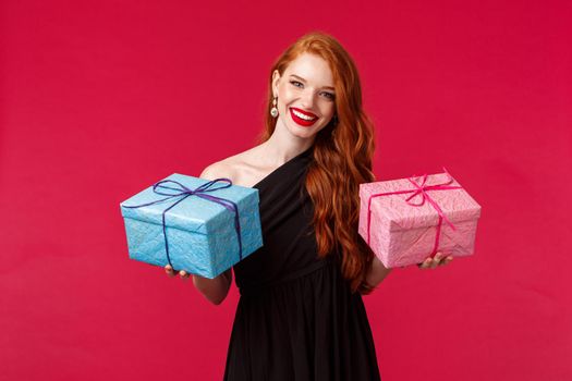 Celebration, holidays and women concept. Portrait of elegant gorgeous young redhead woman in black party dress, holding two gifts in blue and pink boxes, smiling camera cheerful, red background.