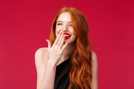 Close-up portrait of pretty elegant young european woman with long curly red hair, black dress and evening makeup, laughing over something funny looking away and cover smile with palm.