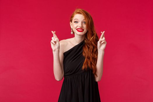 Hopeful and optimistic pretty redhead woman in black evening dress, makeup, cross fingers good luck look at camera pleading eyes, waiting for miracle or dream come true, make wish.