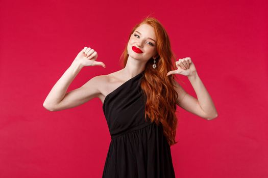 Fashion, luxury and beauty concept. Portrait of sassy and confident, seductive redhead woman in black dress, pointing at herself boasful, bragging and smiling pleased, red background.