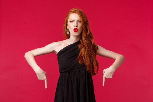 Celebration, events, fashion concept. Portrait of frustrated and upset serious redhead elegant woman in trendy black dress, complaining grimacing dissatisfied, pointing fingers down.